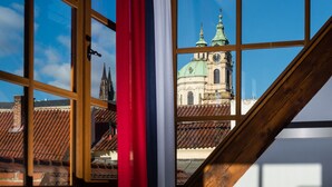 A view through a window with a wooden frame, revealing a building with a green dome and spires, and a red curtain partially visible. - Old Royal Post Boutique Hotel & Premium Suites (Prague)