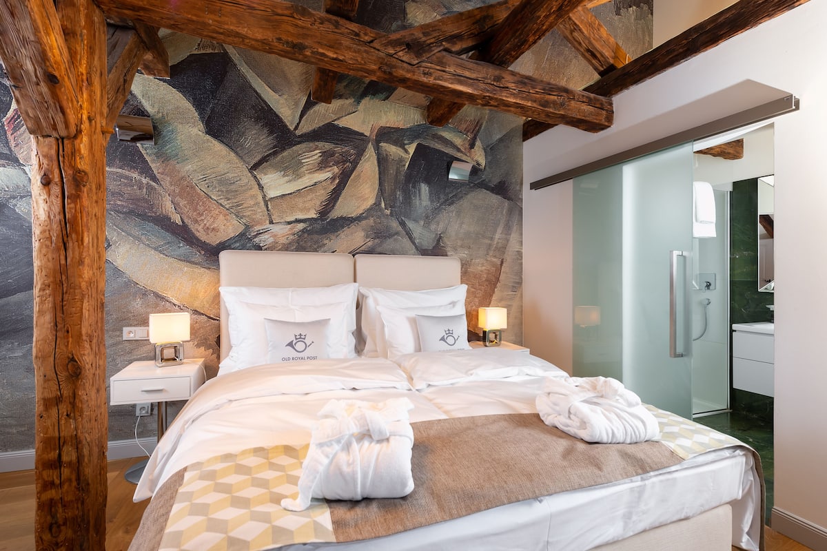 A modern hotel room with a large bed, wooden beams, a textured wall mural, and a glass shower area.