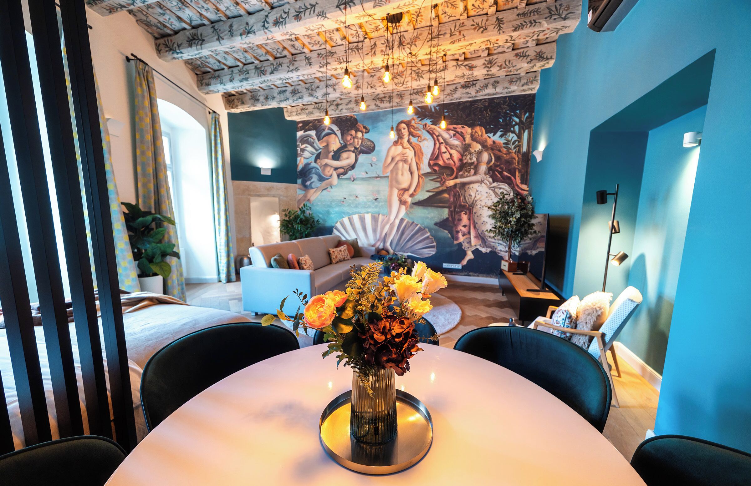 A dining area with a round table, chairs, a vase of flowers, and a mural on the wall.