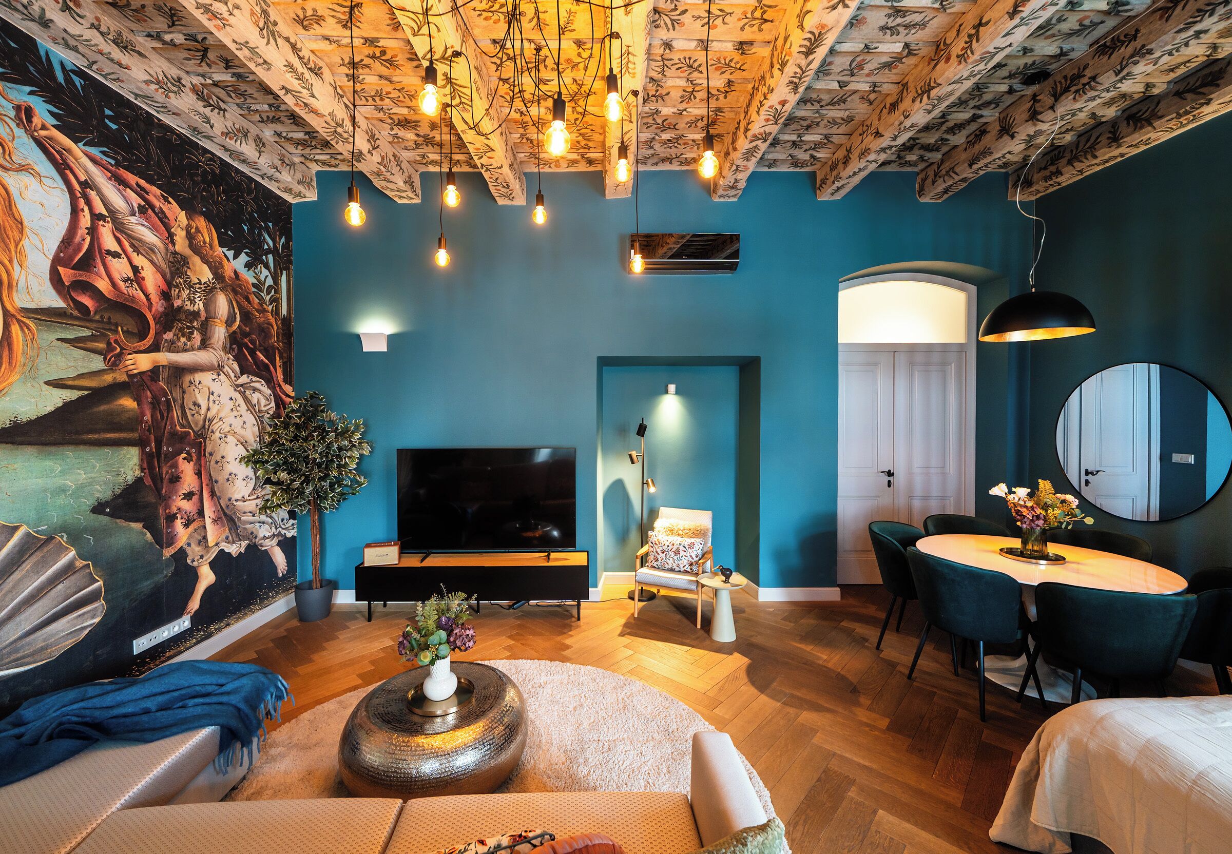 A modern living room with a large mural, a flat-screen TV, a dining area, and a ceiling with exposed beams and patterned wallpaper.
