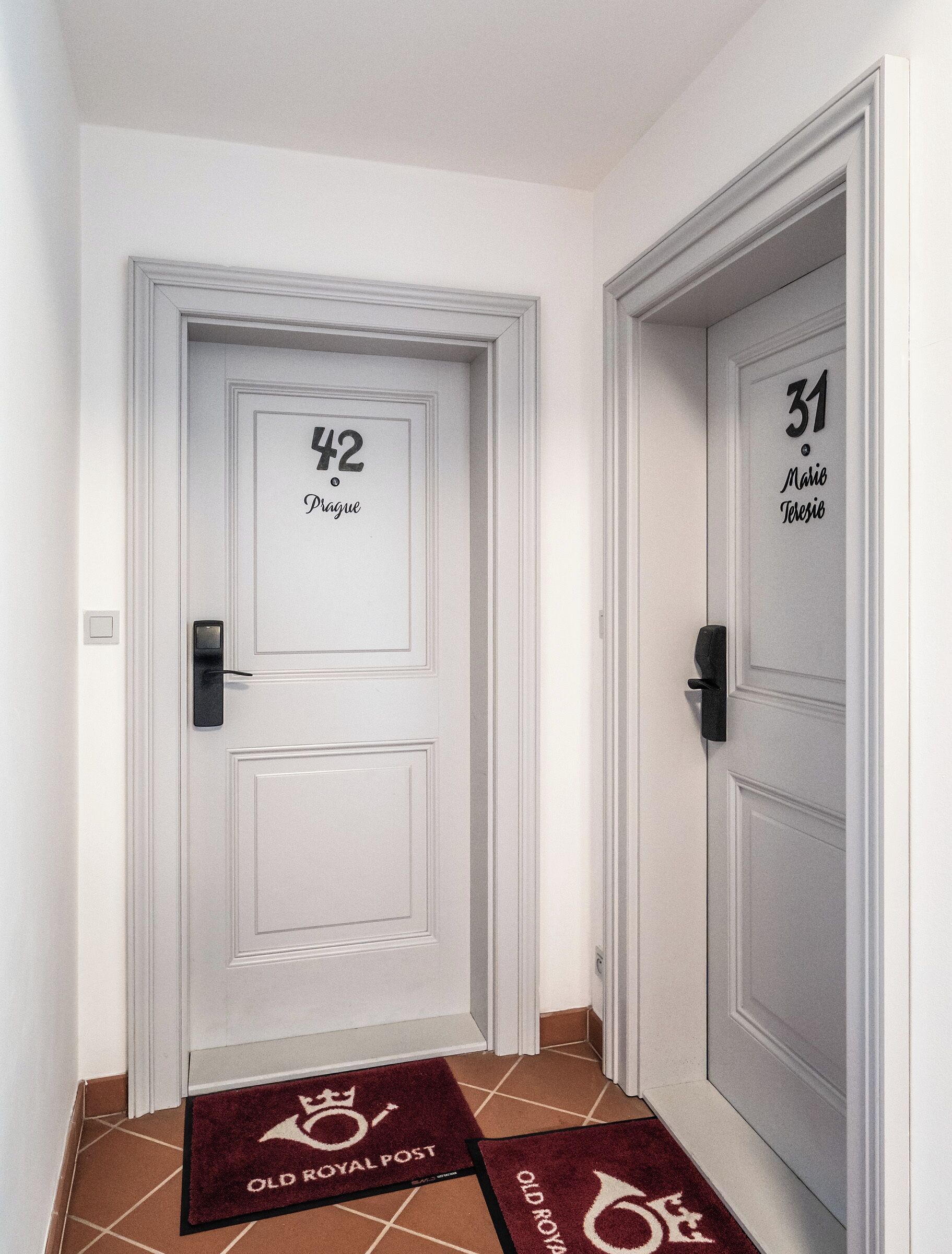 Un couloir avec deux portes, l’une portant l’inscription « 42 Prague » et l’autre « 31 Mario Terezio ». Les deux portes ont des poignées noires et sont peintes en blanc, avec un design simple.