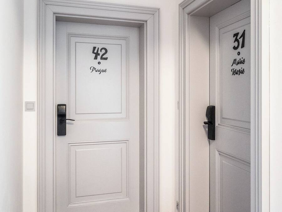 Un couloir avec deux portes, l’une portant l’inscription « 42 Prague » et l’autre « 31 Mario Terezio ». Les deux portes ont des poignées noires et sont peintes en blanc, avec un design simple.