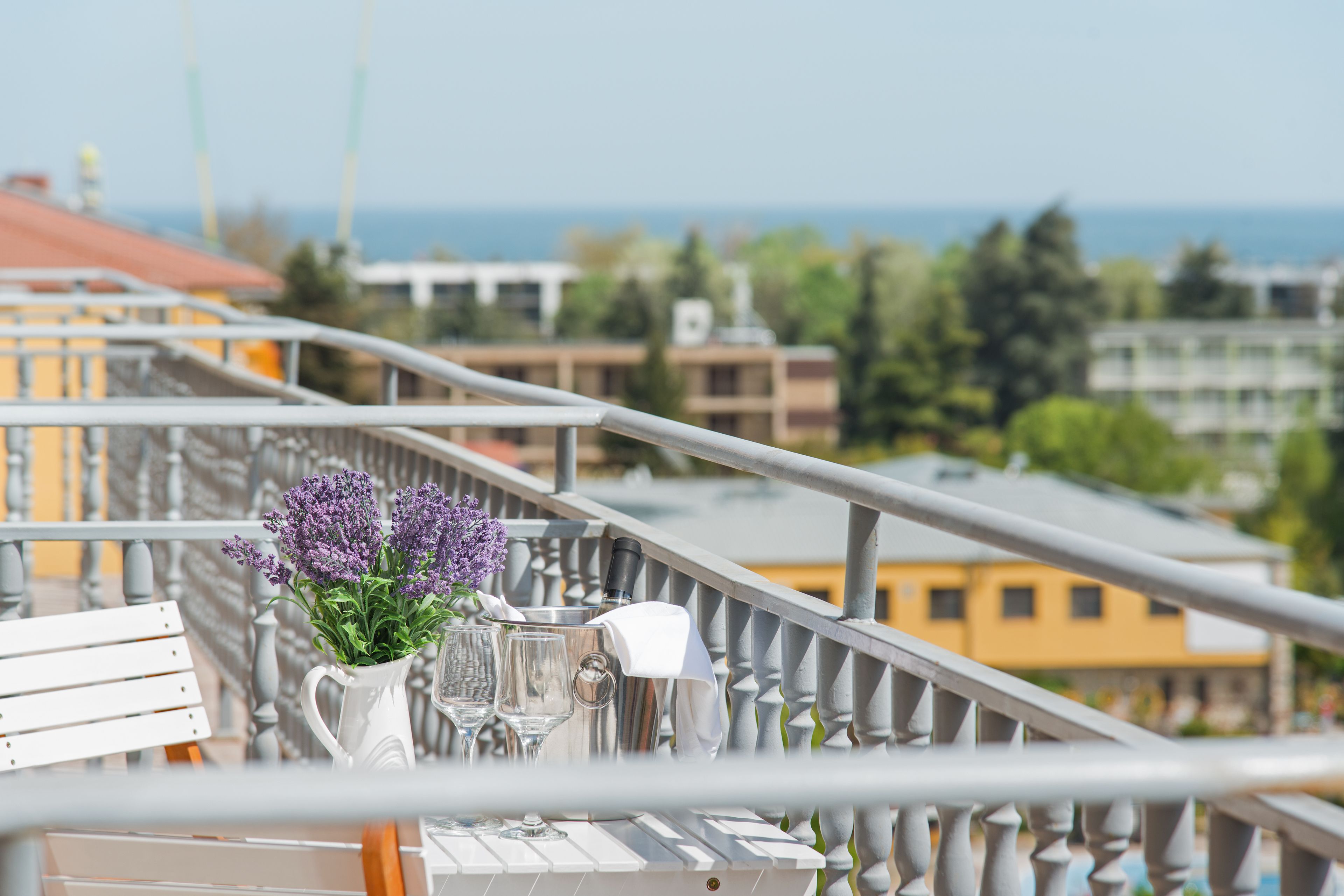 Een balkon met een witte metalen reling, een witte bank en een tafel met een witte karaf, twee wijnglazen en een bos paarse bloemen.