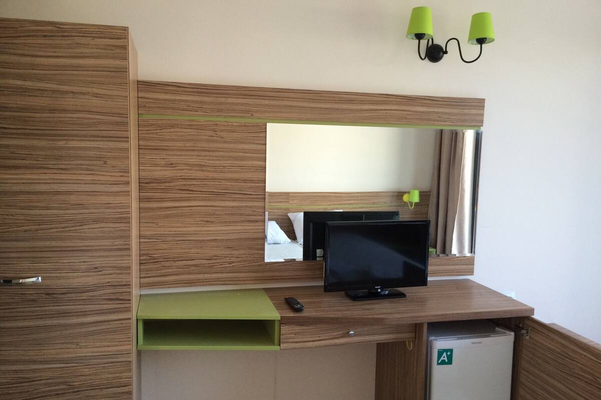 A hotel room with a wooden desk, a television, a green shelf, a small refrigerator, and a mirror above the desk.