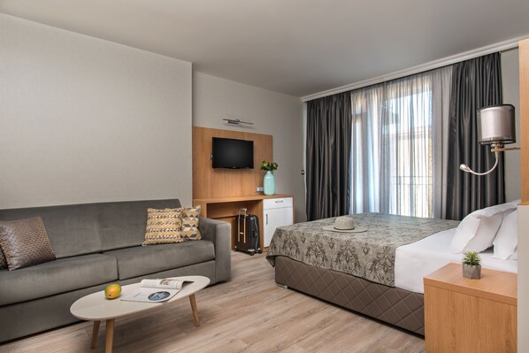 A modern hotel room with a sofa, a bed, a desk, a television, and a window with curtains.