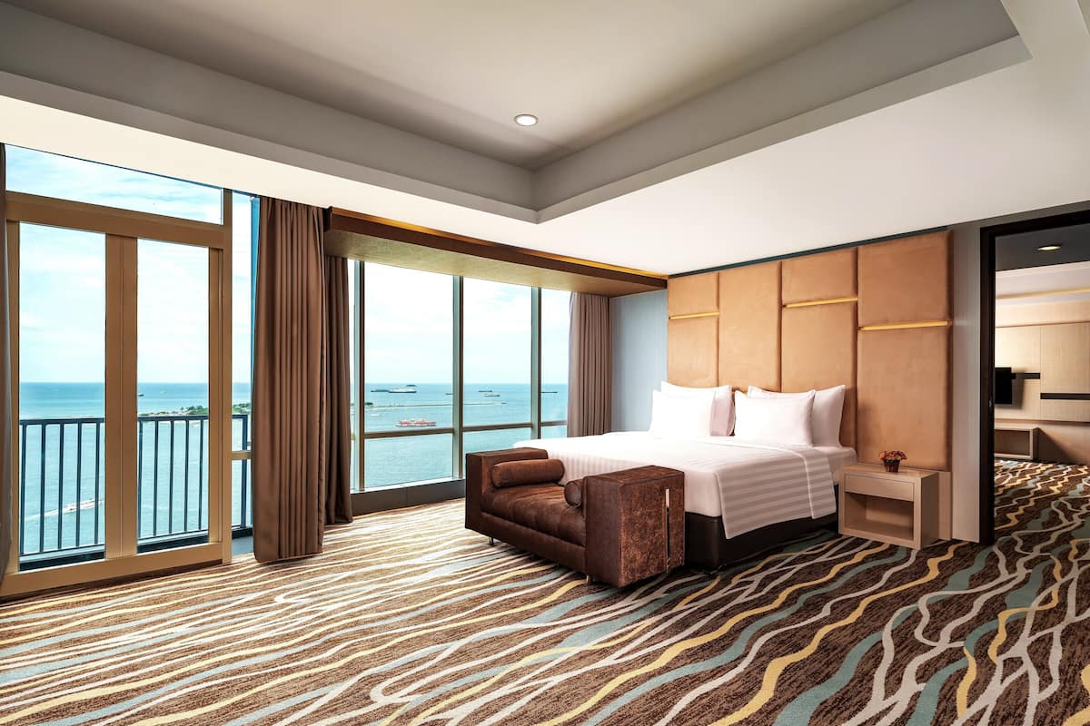 Habitación de hotel con una cama grande, un sofá, una mesita de noche y vistas al mar a través de amplias ventanas.