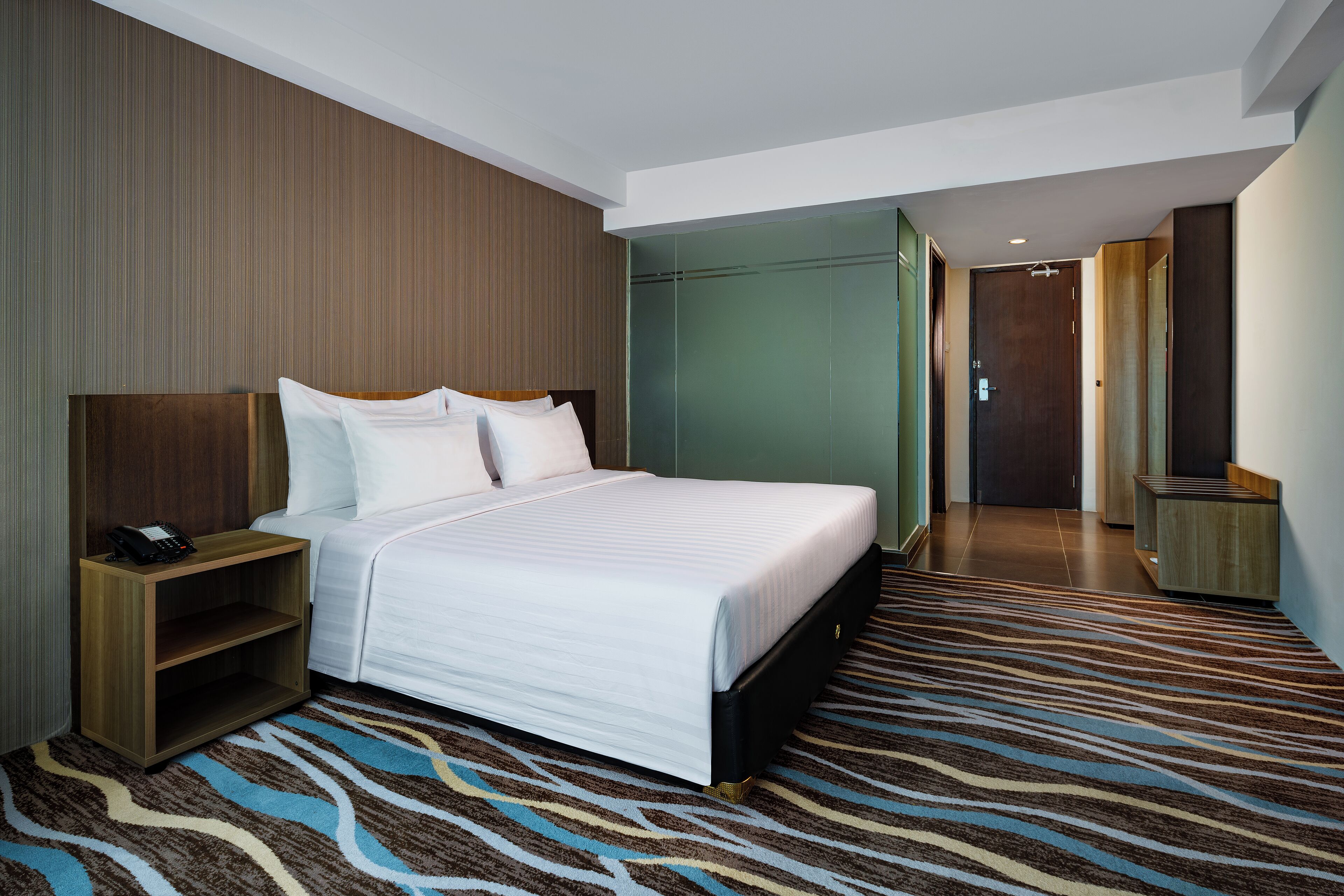 A modern hotel room with a large bed, a bedside table, a telephone, and a patterned carpet.