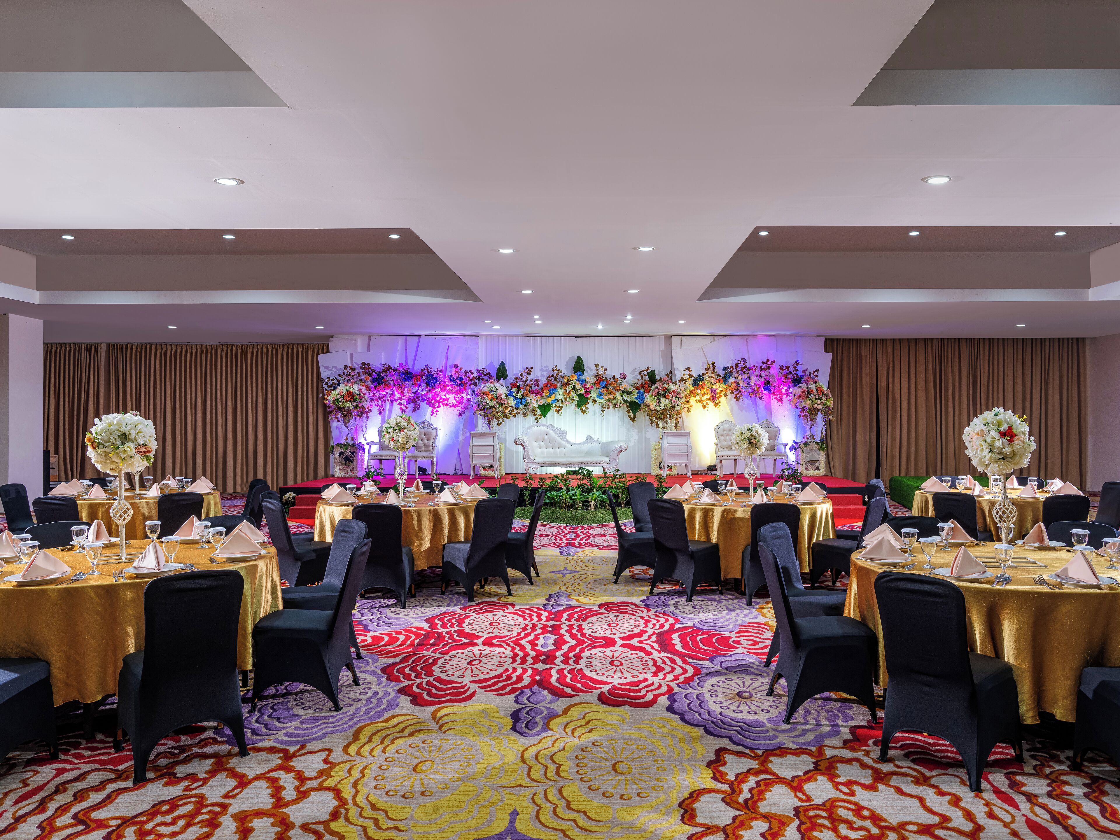 A banquet hall with round tables, black chairs, and a stage with floral decorations.