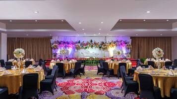 A banquet hall with round tables, black chairs, and a stage with floral decorations.