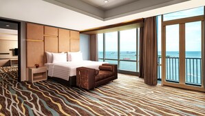 Une chambre d’hôtel avec un grand lit, un canapé, une table de chevet et une vue sur la mer grâce à de grandes fenêtres.