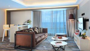 Une chambre d’hôtel moderne avec un canapé marron, un bureau blanc, une table basse ronde et un téléviseur fixé au mur.