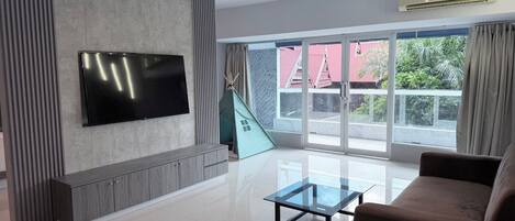 Un soggiorno moderno con televisore a schermo piatto montato a parete, un tavolino da caffè in vetro, un divano marrone e un'ampia porta scorrevole in vetro che si apre su un balcone con vista su vegetazione e un edificio con tetto rosso.
