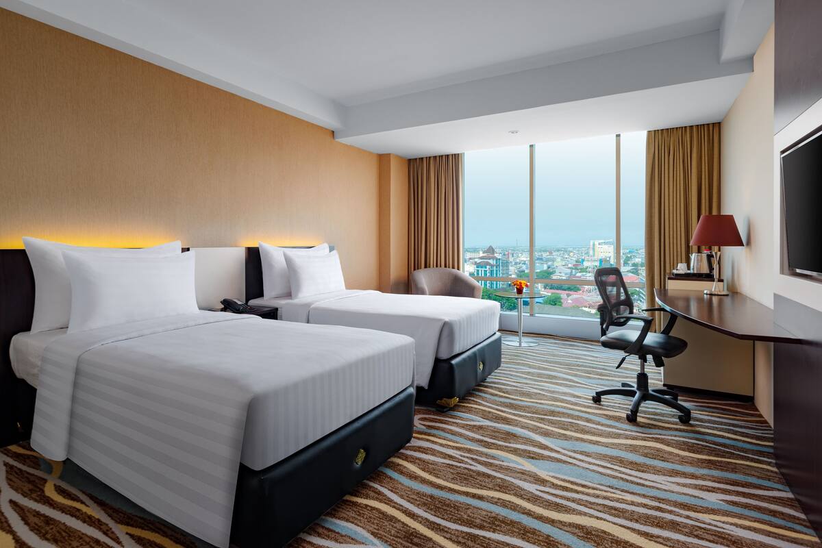 Habitación de hotel con dos camas, un escritorio, una silla y un ventanal con vistas a la ciudad.