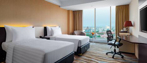 A hotel room with two beds, a desk, a chair, and a large window offering a city view.