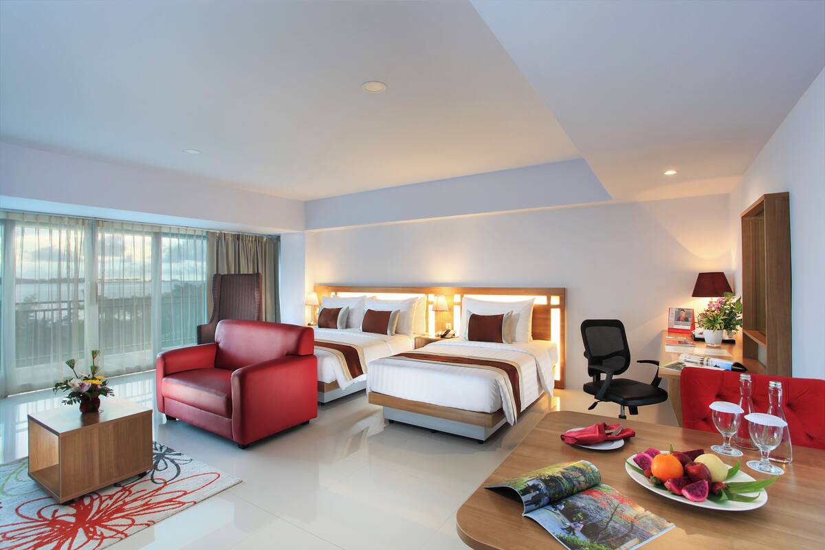 Habitación de hotel con dos camas, un sillón rojo, una mesita de madera, una mesa de comedor con una bandeja de frutas y un escritorio con una silla.