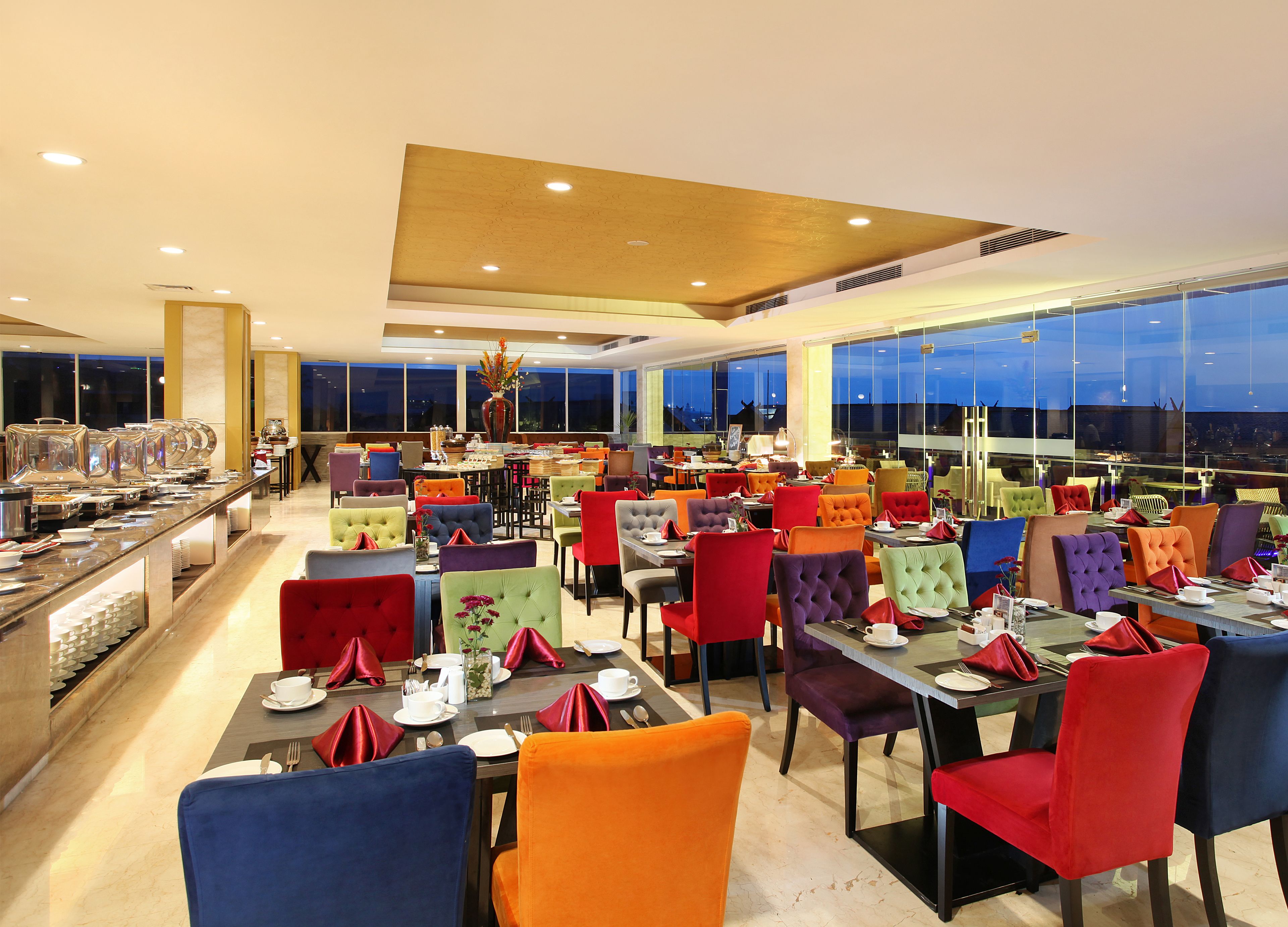 Un comedor amplio con múltiples mesas y sillas, ventanas grandes que ofrecen vista del paisaje urbano al atardecer, y una barra de buffet con diversos alimentos.