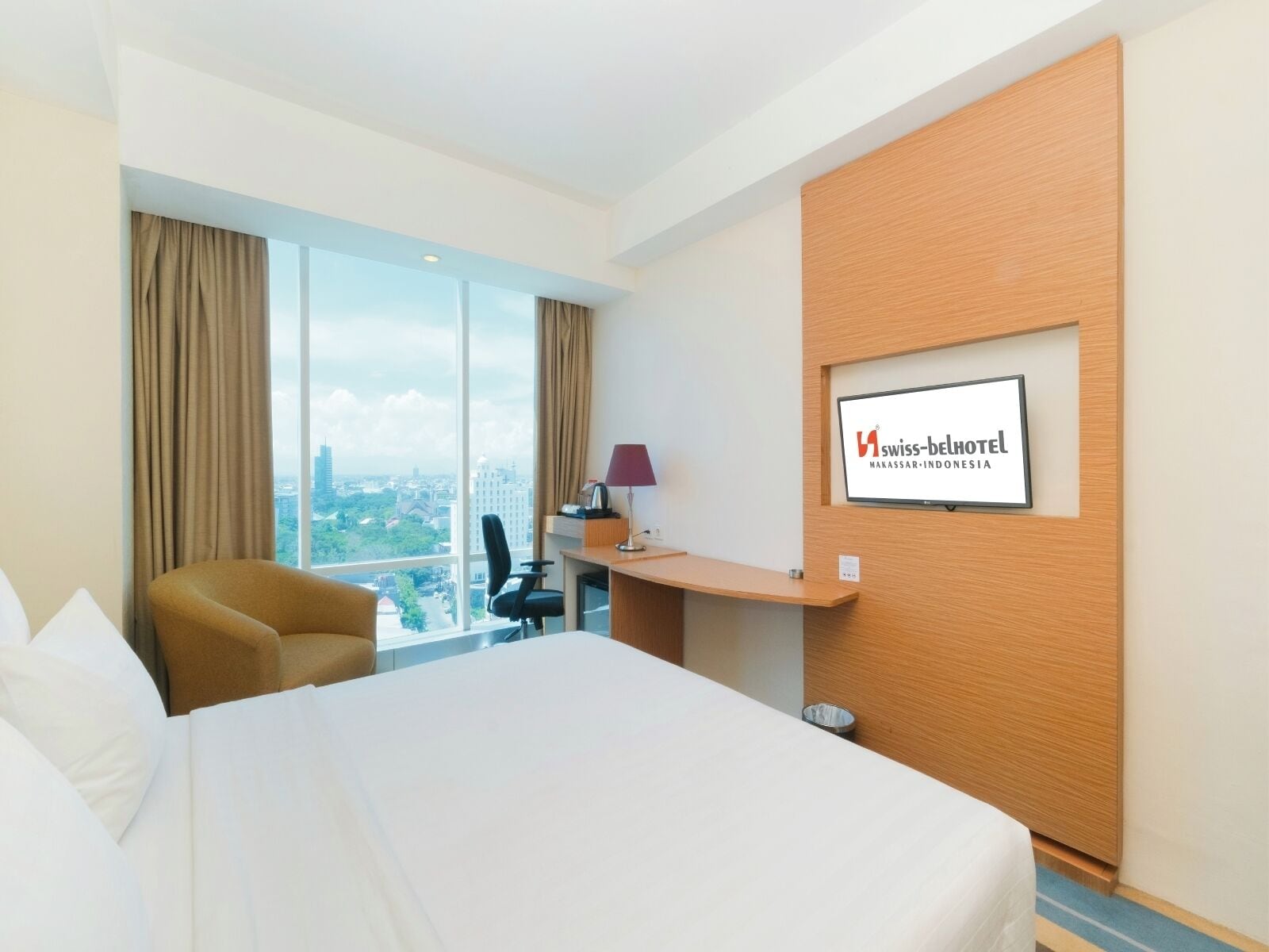 A hotel room with a large bed, a desk, a chair, and a TV displaying the hotel name.