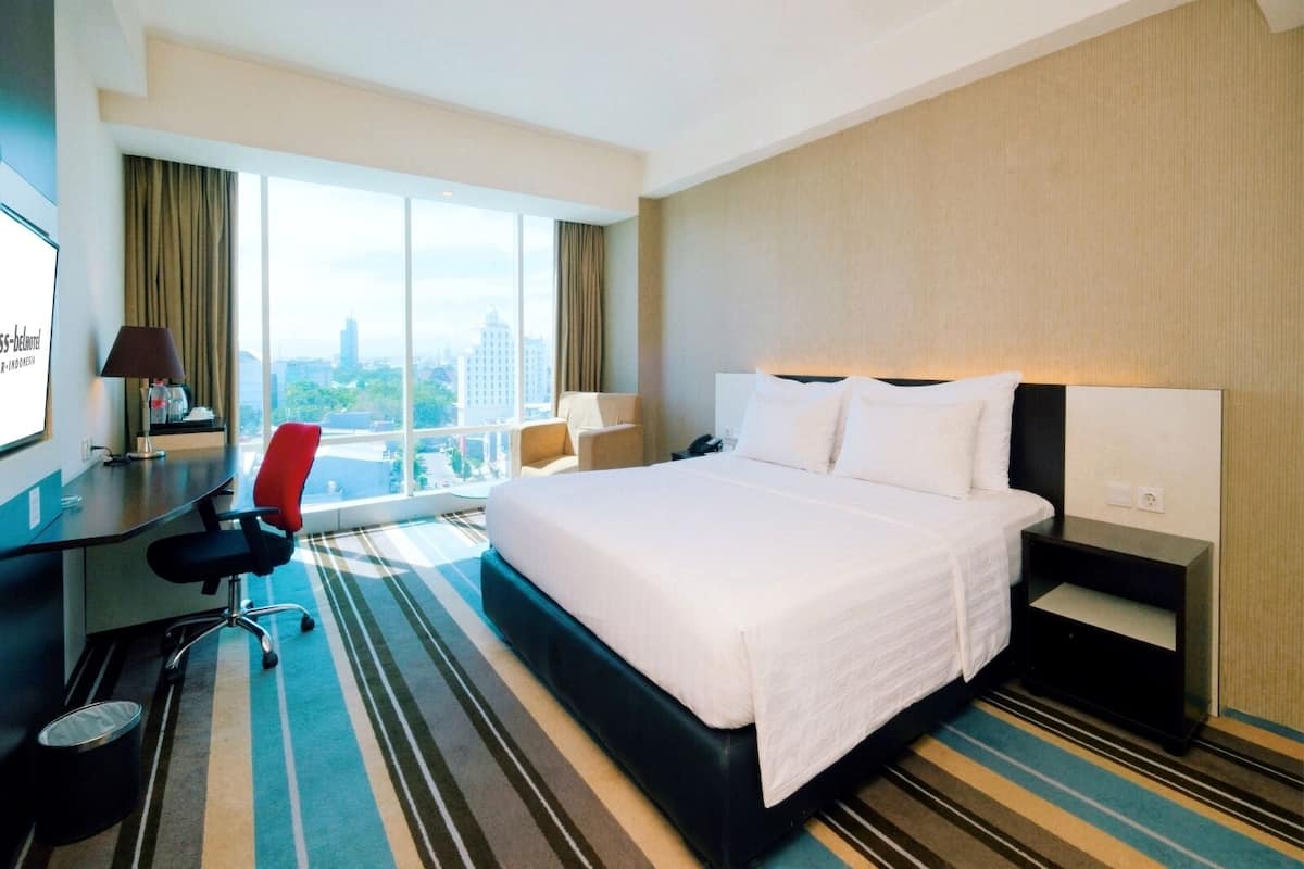 Habitación de hotel con una cama grande, un escritorio con una silla, una lámpara y vistas de la ciudad a través de amplios ventanales.