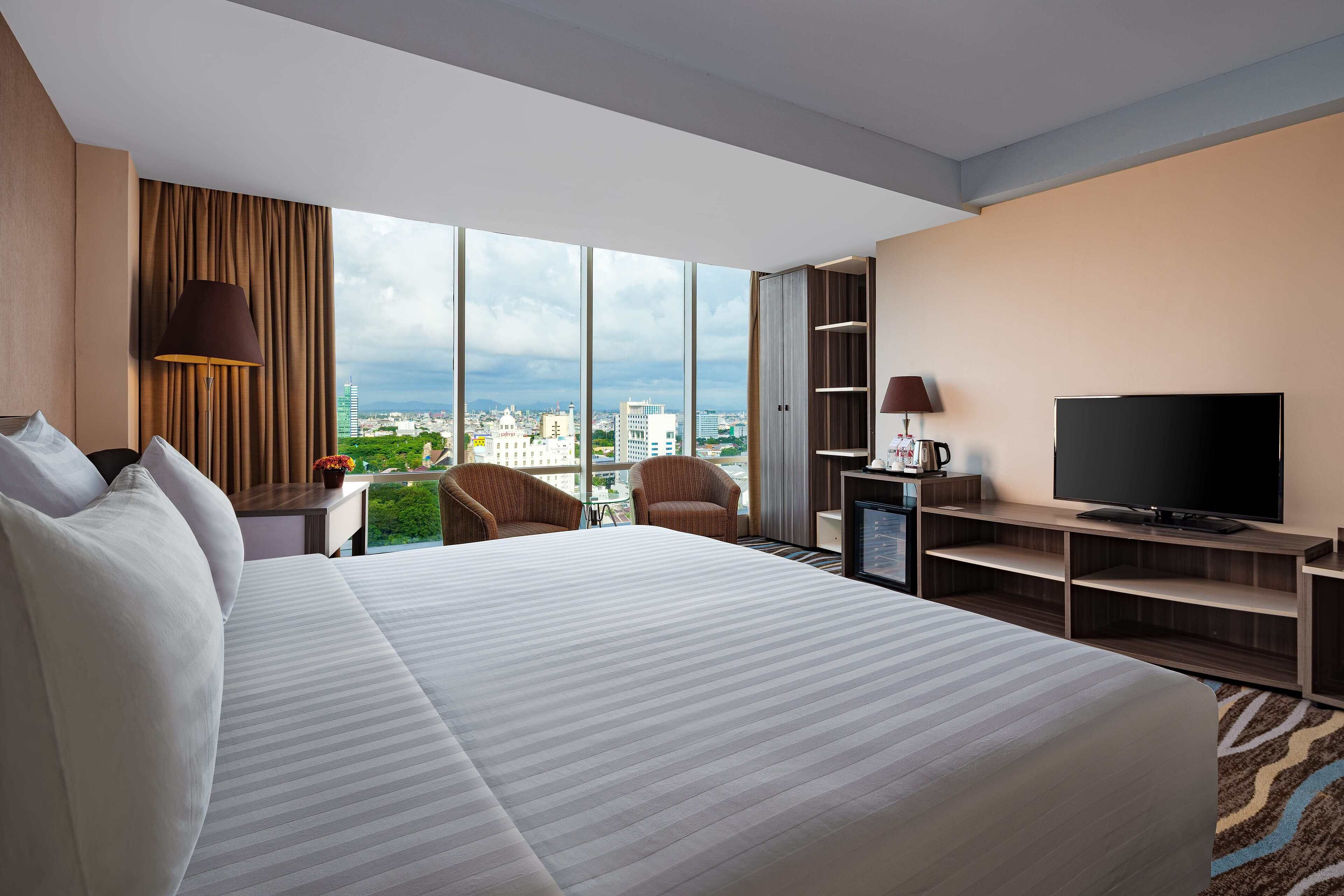 A hotel room with a large bed, two armchairs, a TV, and a view of the city.