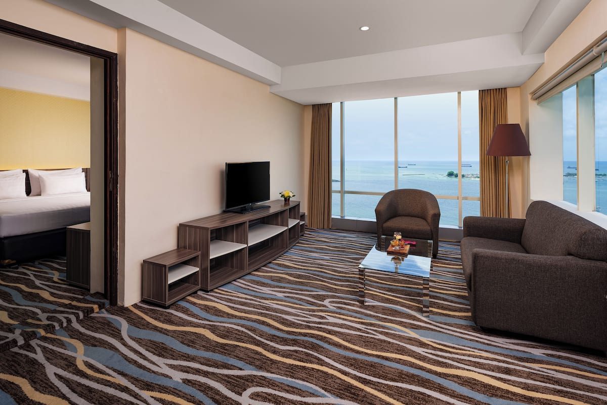 Habitación de hotel con cama, televisión, sofá, una mesa de centro y un gran ventanal con vista al mar.