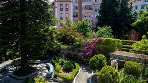 Un jardín ajardinado con diversas plantas y árboles, una fuente y un edificio con balcones al fondo.