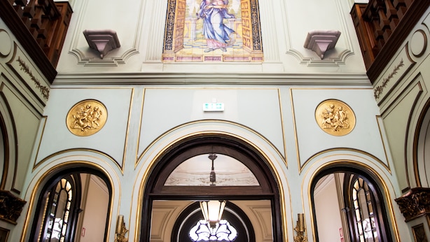Fachada ornamentada de un edificio con un ventanal central de vidrieras que representa una figura, flanqueado por dos medallas doradas y un amplio arco de entrada.