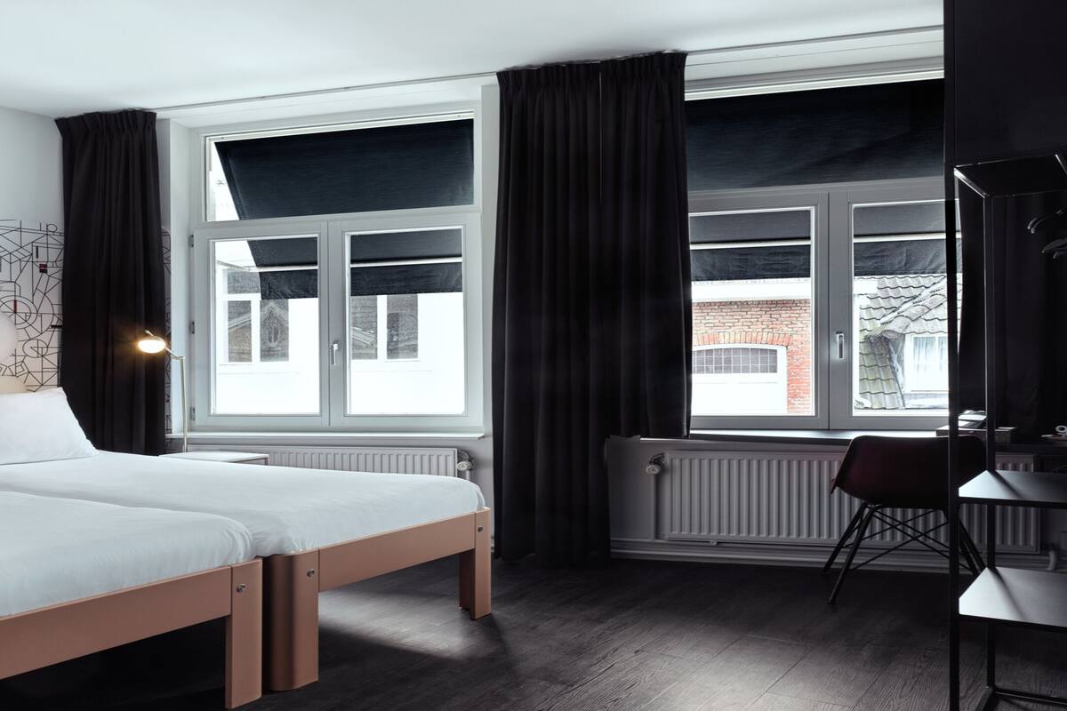 A modern hotel room with a large window, a bed, a desk, and a radiator.