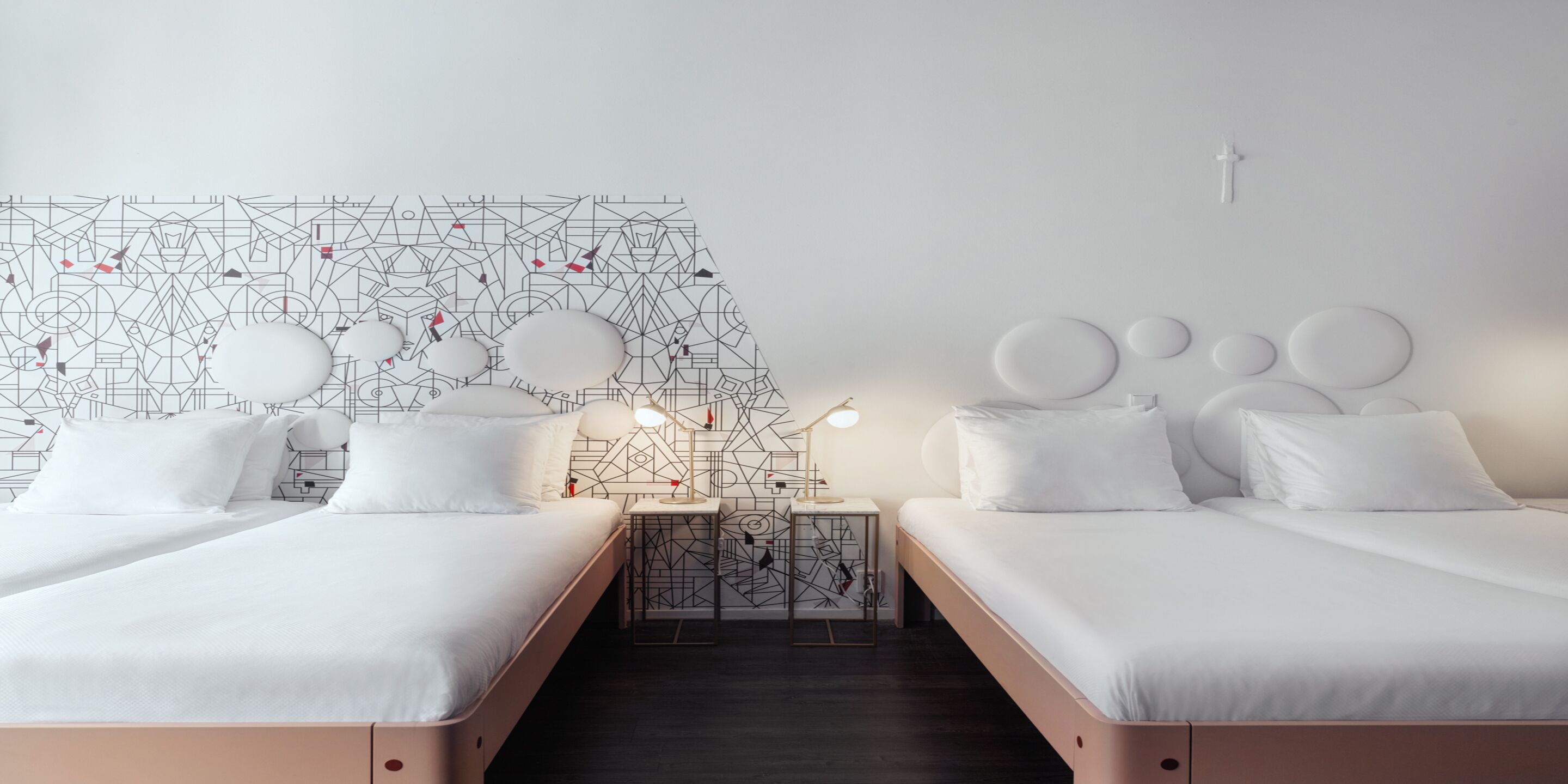 Una habitación de hotel moderna con dos camas individuales, una pared con diseño geométrico, una mesita de noche pequeña con lámpara y una decoración minimalista.