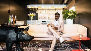 A person sitting on a white sofa in a modern interior with a geometric patterned carpet, a black pig sculpture, and a bar area with shelves of bottles and glasses in the background. - MABI City Centre (Maastricht)