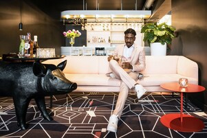 A person sitting on a white sofa in a modern interior with a geometric patterned carpet, a black pig sculpture, and a bar area with shelves of bottles and glasses in the background.