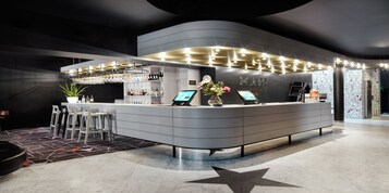A modern hotel reception area with a curved counter, digital check-in kiosks, and a star-patterned floor.