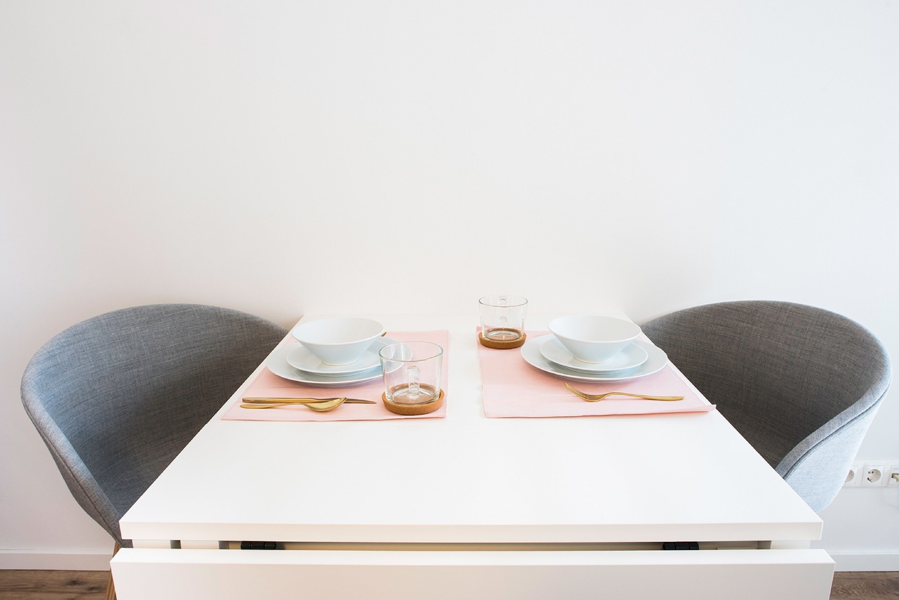 A neatly set dining table with two chairs, white table, pink placemats, and gold cutlery.