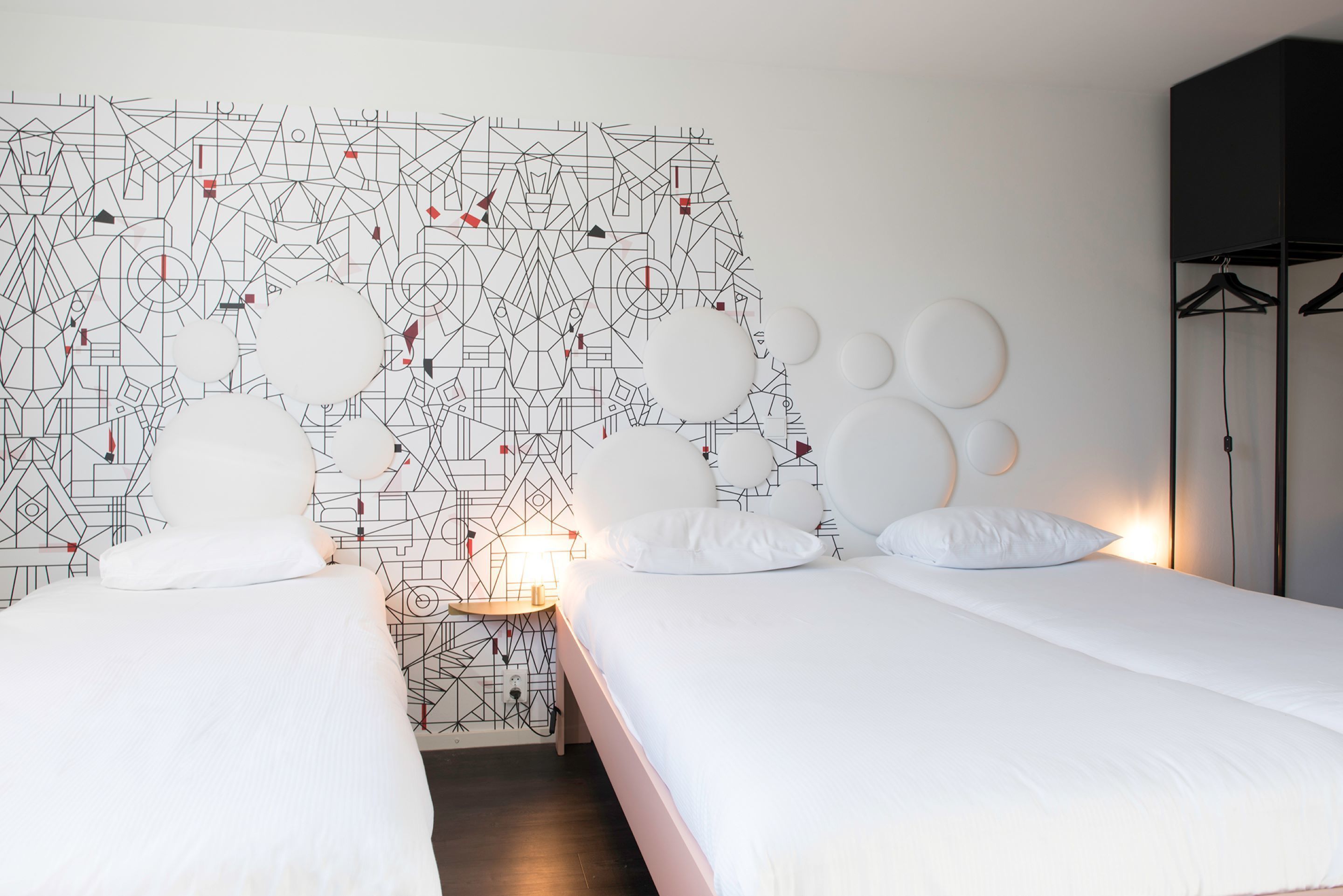 Habitación de hotel con dos camas, un muro con estampado geométrico, una mesa redonda con lámpara y un armario negro.
