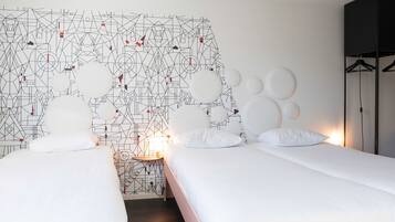 Habitación de hotel con dos camas, un muro con estampado geométrico, una mesa redonda con lámpara y un armario negro.