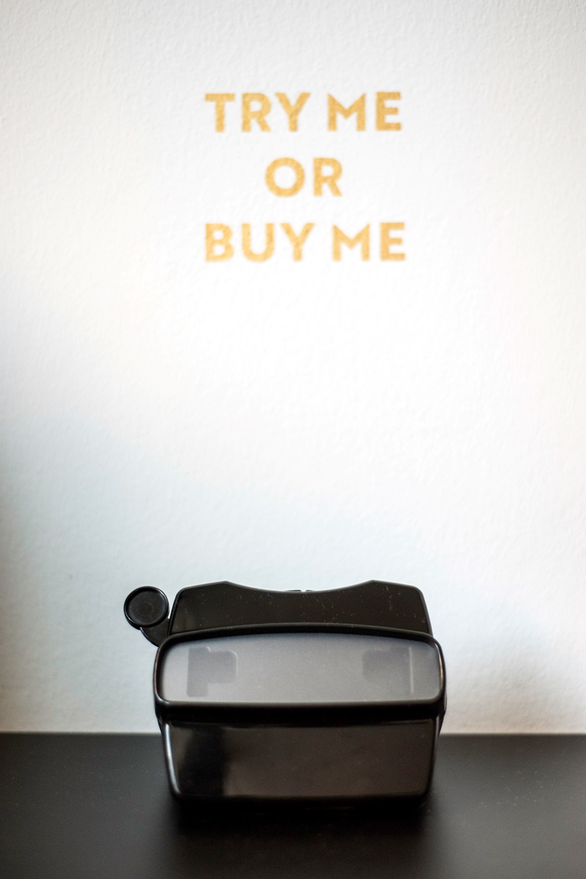 Une trousse de toilette noire avec une face transparente et une fermeture éclair, posée sur une surface noire devant un mur blanc, et sur laquelle on peut lire le texte : « Essayez-moi ou achetez-moi ».