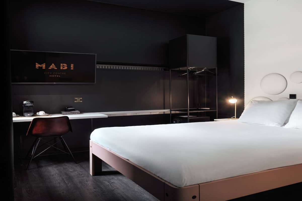 A modern hotel room with a bed, desk, chair, and a wall-mounted TV displaying the hotel name.