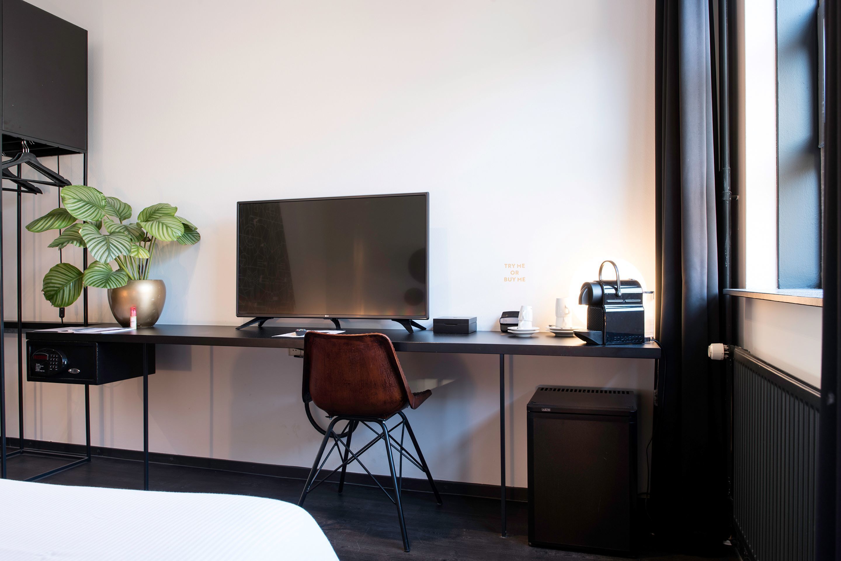 Een moderne hotelkamer met een bureau, een flatscreen televisie, een stoel, een potplant, een radiator en een prullenbak.