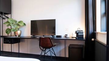 Een moderne hotelkamer met een bureau, een flatscreen televisie, een stoel, een potplant, een radiator en een prullenbak.
