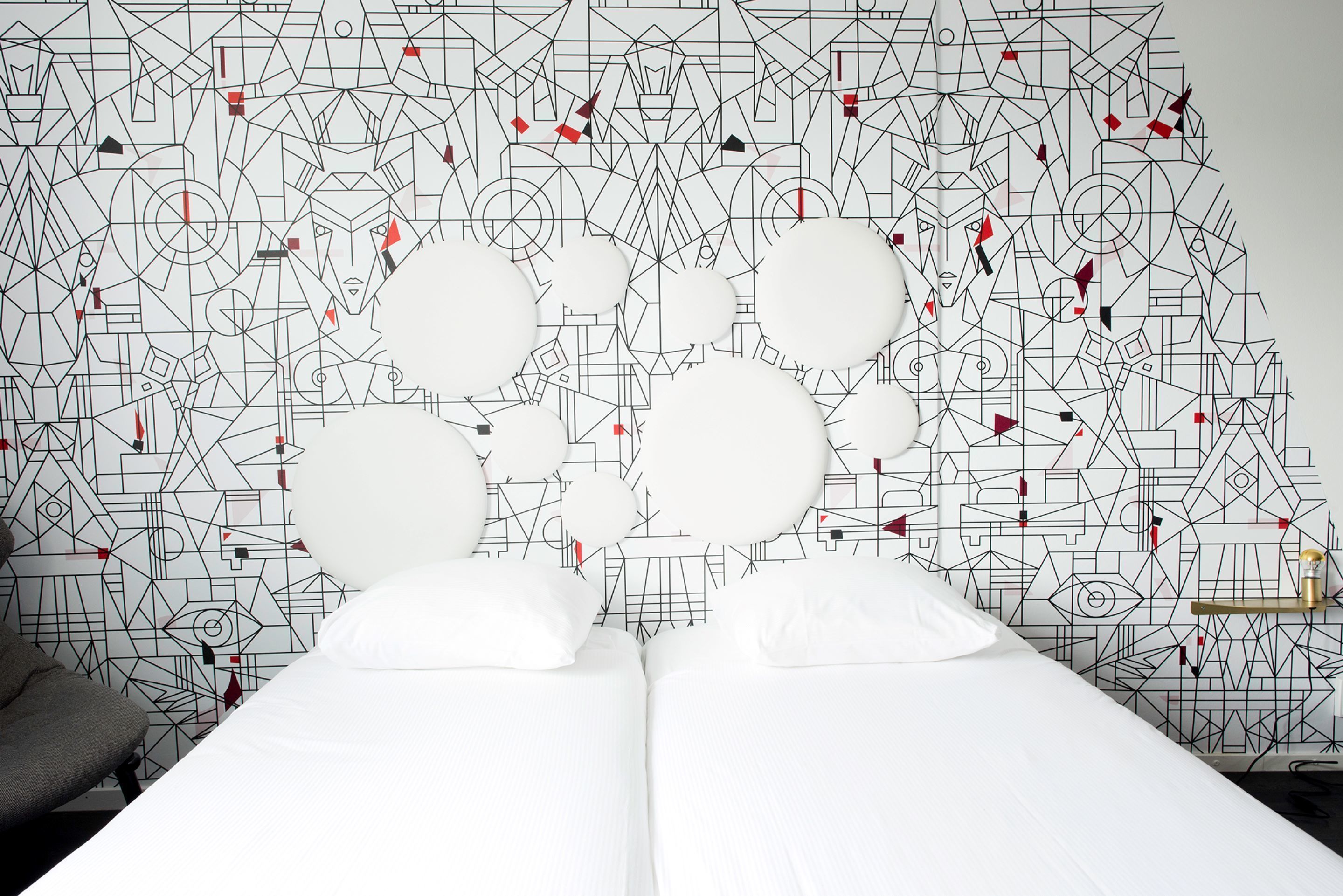 Een hotelkamer met behang met een geometrisch patroon, een bed met witte lakens en een nachtkastje met een lamp.