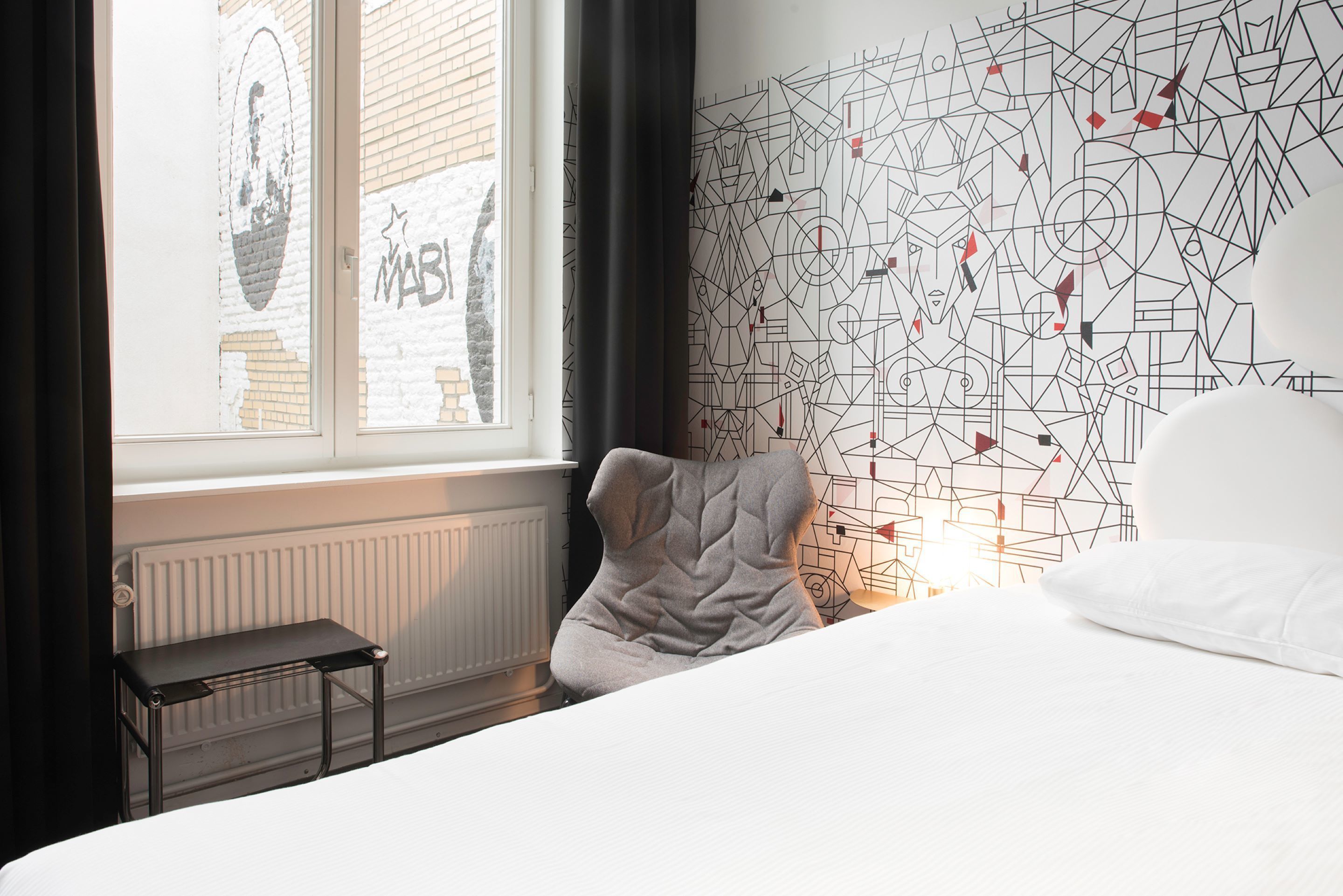 A bedroom with a bed, a window, a radiator, and a wall with geometric patterns.