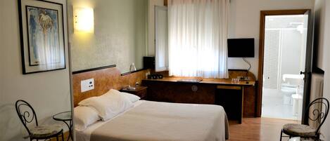 A hotel room with a bed, a desk, a chair, a TV, and a bathroom visible through an open door.
