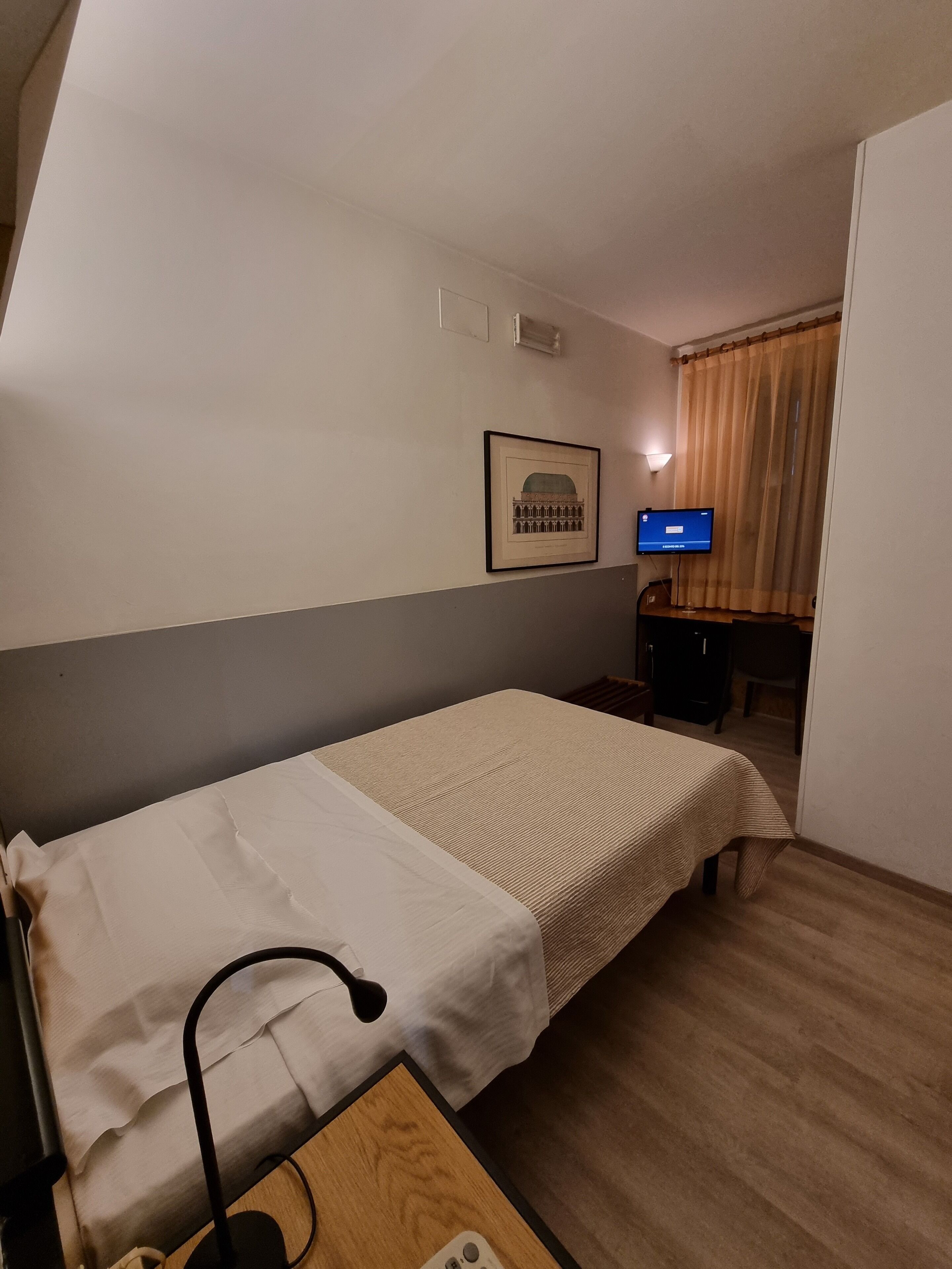 A small hotel room with a single bed, a desk with a lamp, a television, and a framed picture on the wall.