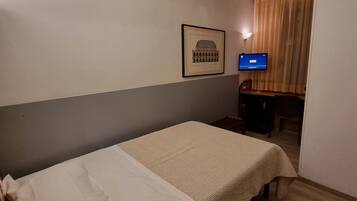 A small hotel room with a single bed, a desk with a lamp, a television, and a framed picture on the wall.