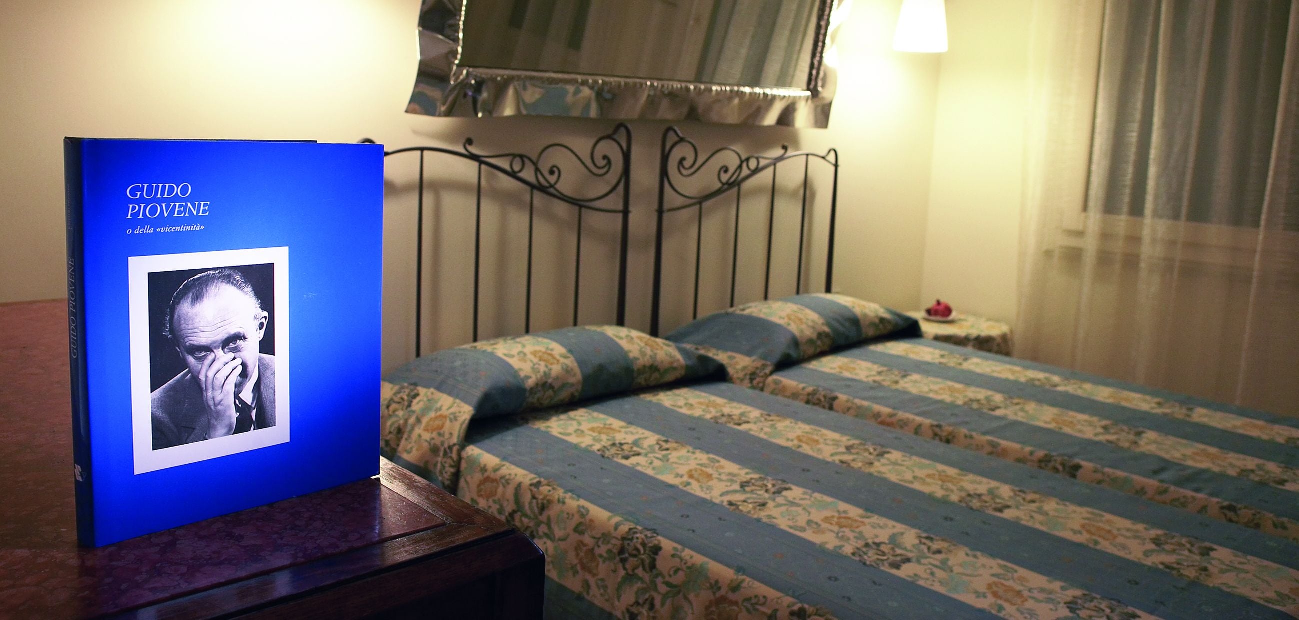 Een hotelkamer met een bed met gestreept beddengoed, een nachtkastje met een ingelijste foto en een raam met gordijnen.