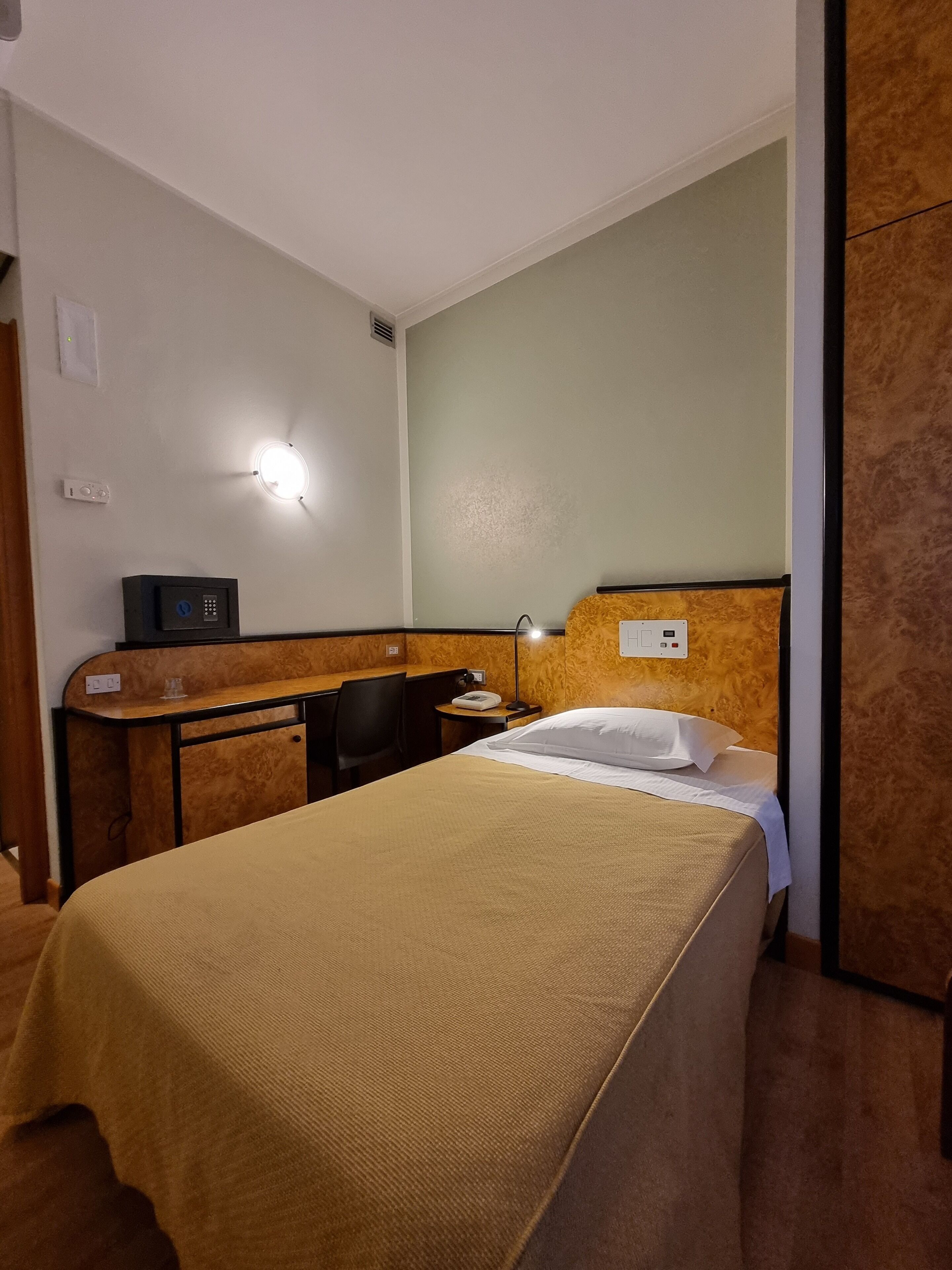 A hotel room with a bed, a desk, a chair, a lamp, and a wall-mounted control panel.