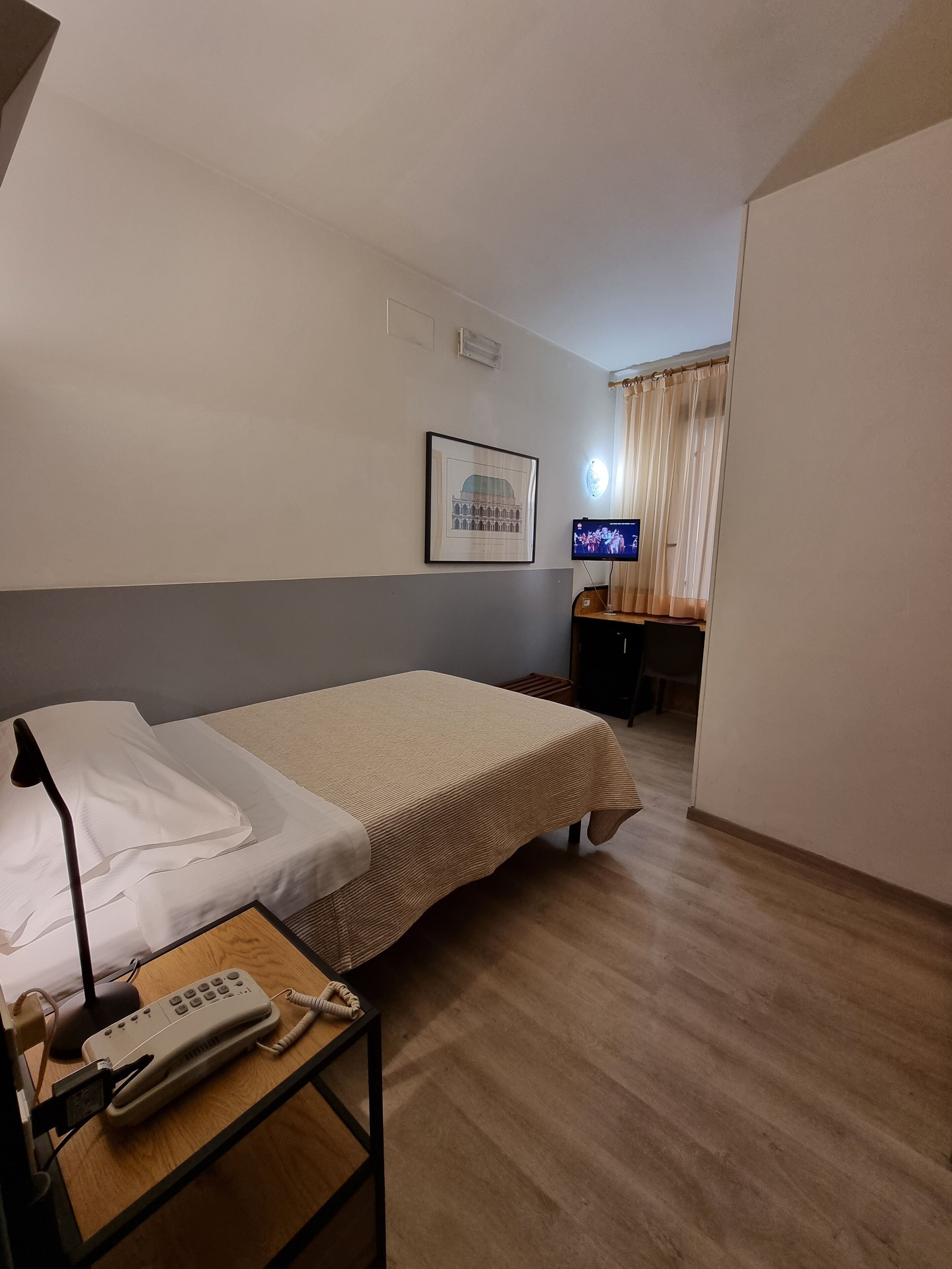 Een hotelkamer met een bed, een bureau met een computer, een ingelijste foto aan de muur en een telefoon op een klein tafeltje.