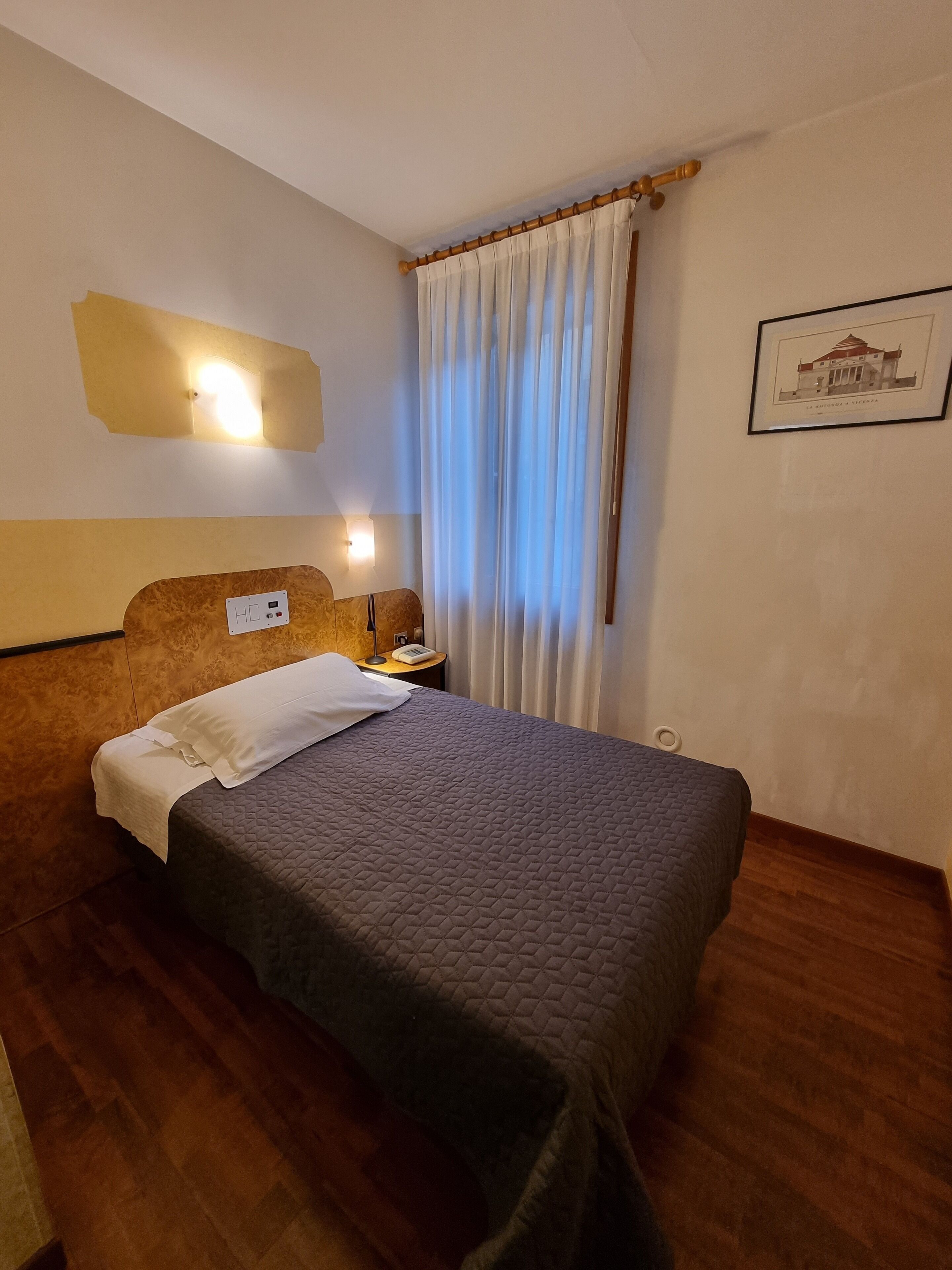 Una habitación de hotel con cama, mesita de noche, lámpara y un cuadro en la pared.