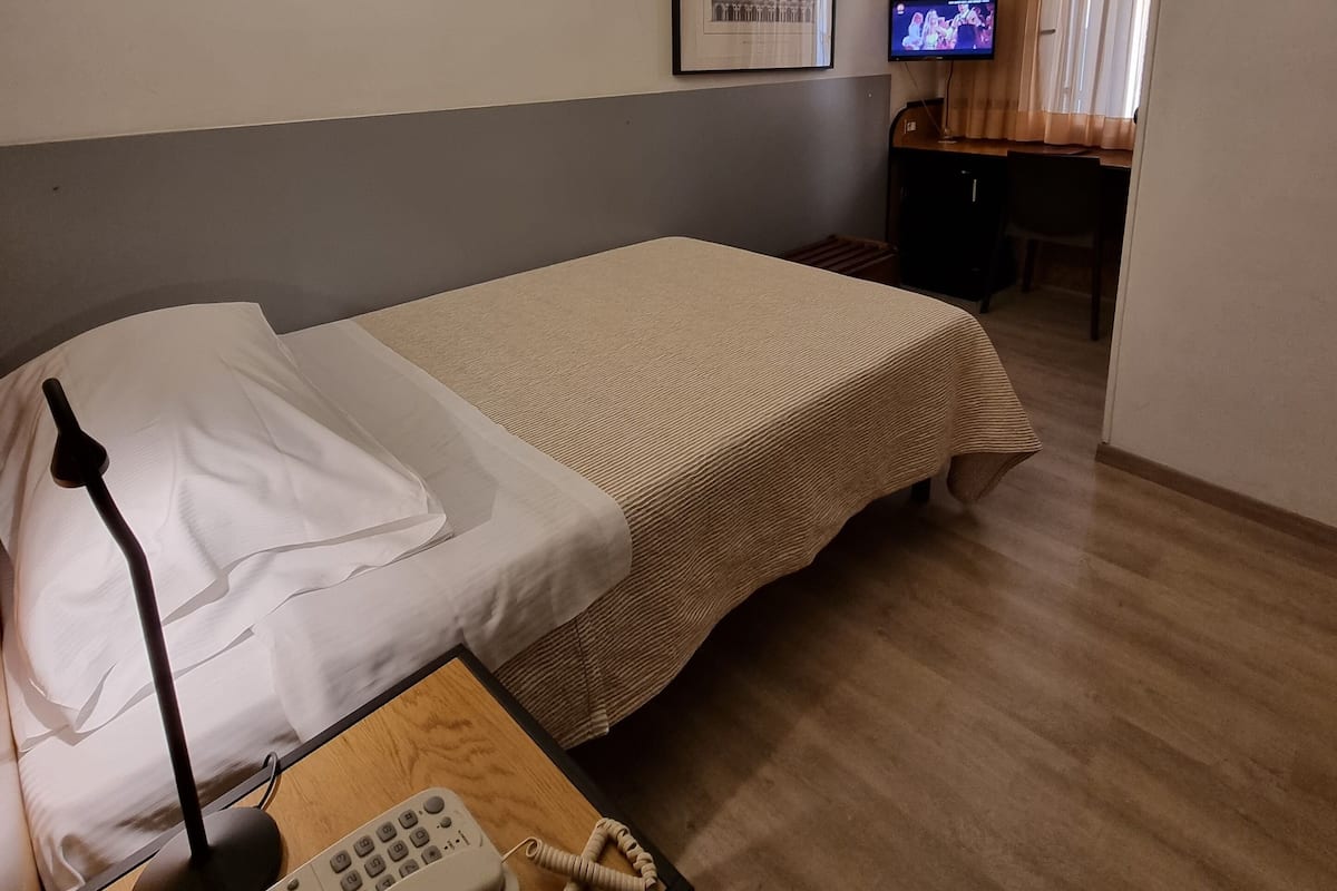 Habitación de hotel con cama, mesita de noche con teléfono y lámpara, televisor en un escritorio y un cuadro en la pared.