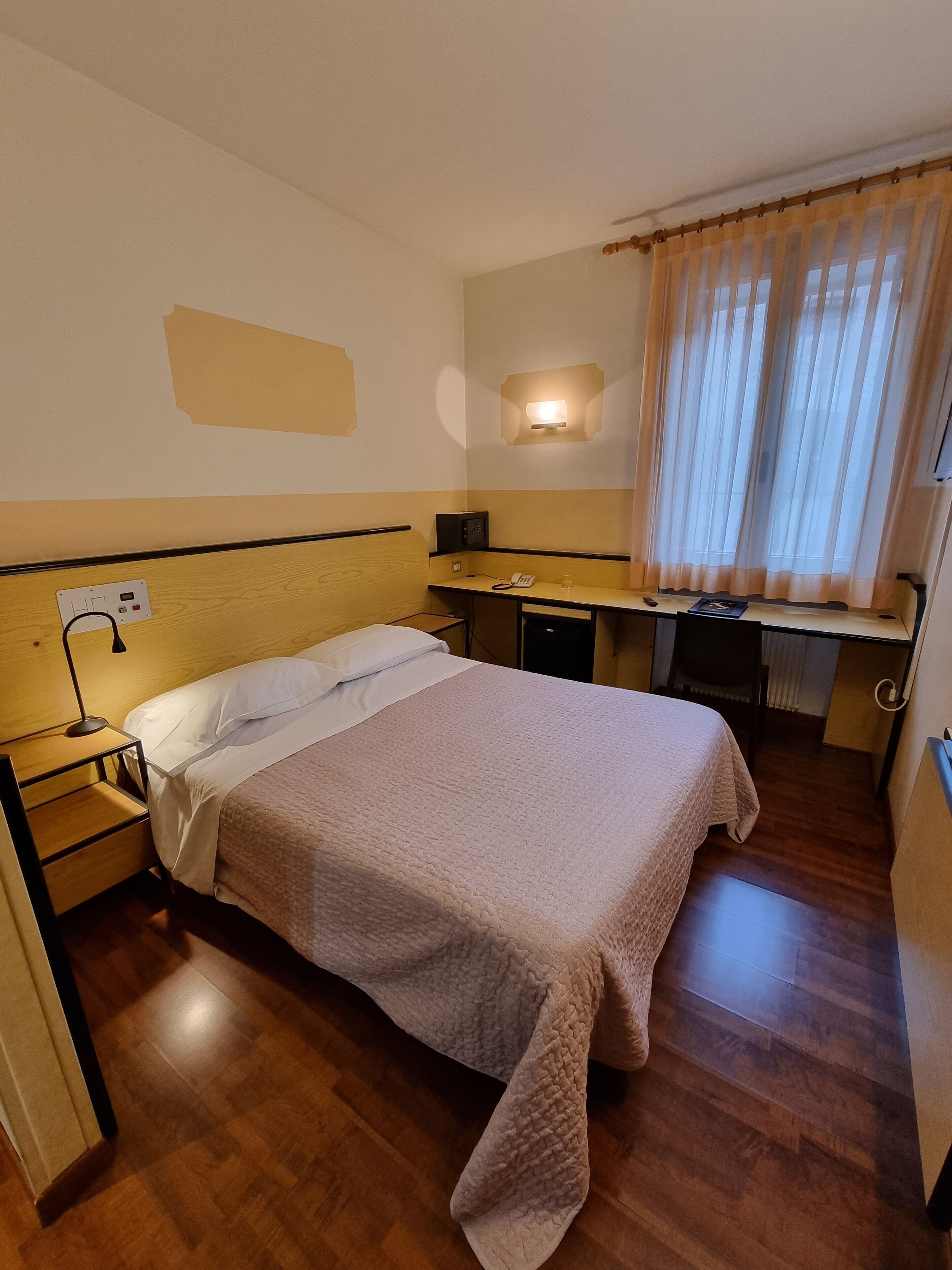 A hotel room with a bed, bedside table, desk, and a window with curtains.