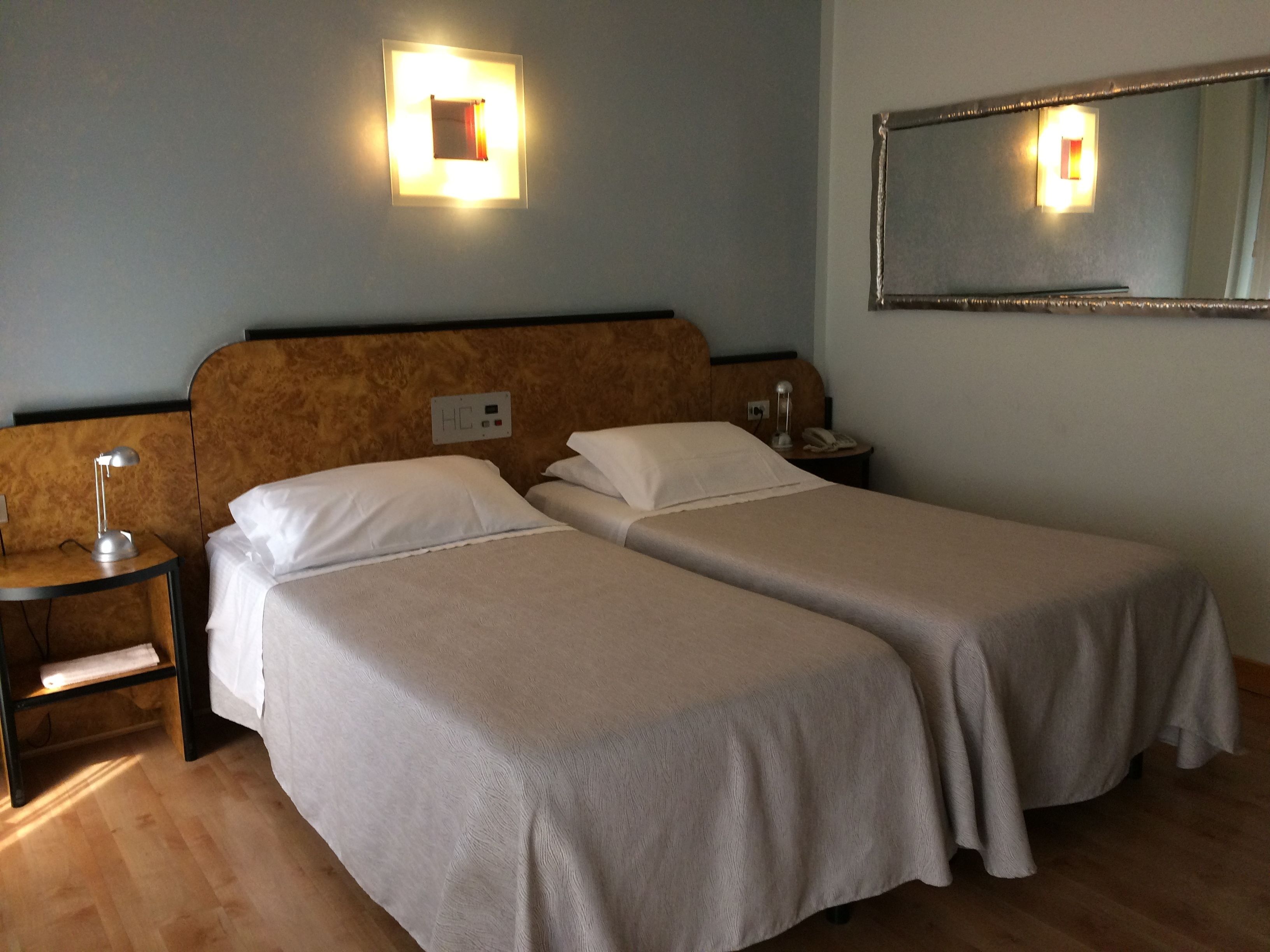 A hotel room with two single beds, a nightstand with a lamp, a bedside table with a phone, and a wall-mounted light fixture.